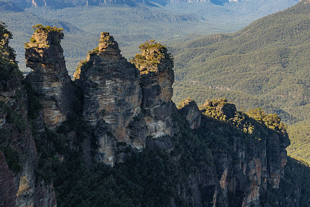 The Three Sisters, a rock formation in the Blue Mountains of New South Wales, Australia, stand tall against a backdrop of lush green forest.