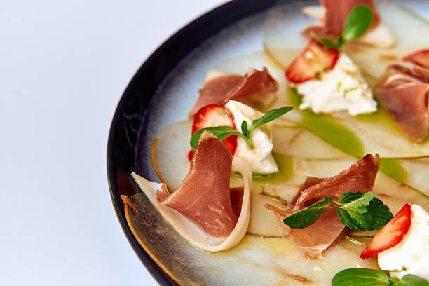 istock A gourmet plate featuring thinly sliced prosciutto arranged elegantly with fresh strawberries, creamy cheese, and a drizzle of vibrant sauce on a modern dish. 2227086107