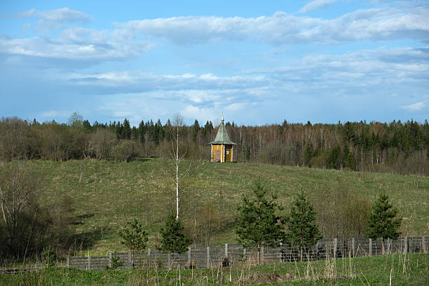 A charming wooden chapel rests on a grassy hill amidst a beautiful landscape of trees under a bright blue sky. The setting highlights nature's tranquility.