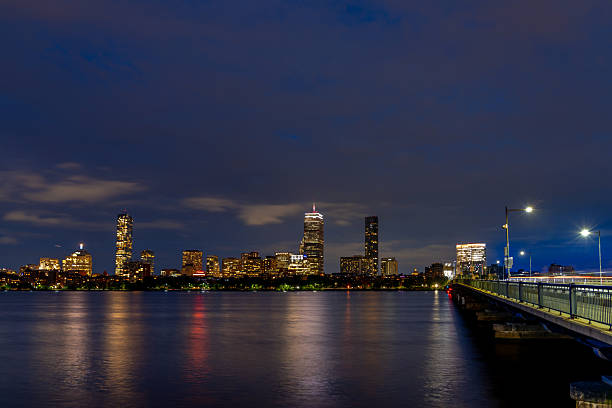 Boston's skyline glows at night, seen from a bridge over the Charles River. City lights reflect on the water.