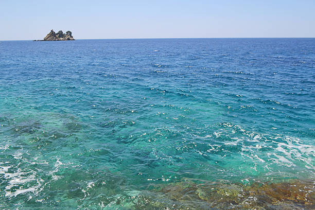 Traditional stone buildings with charming shutters and lush green vines under a clear blue sky in Petrovac, Montenegro. Dramatic layered rock formations and lush green trees meet the vibrant turquoise and blue waters of the Adriatic Sea.