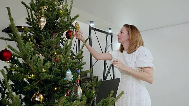 Female decorating christmas tree with golden ornament, smiling while creating festive holiday ambiance in cozy home interior
