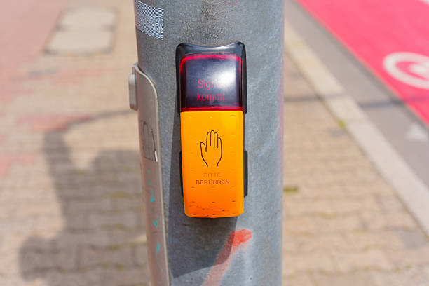 Close-up view of a pedestrian traffic signal button on a metal pole, featuring warning lights and instructions in German, set against a city street background.