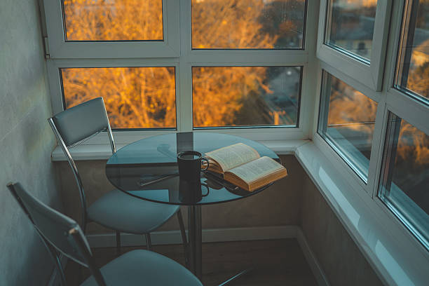 Cozy reading nook with an open book and black cup on a round glass table by a panoramic window, overlooking autumn trees glowing in golden sunset light