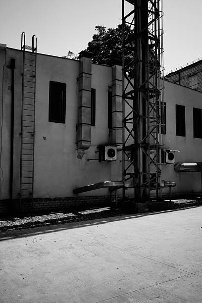 A black and white image capturing the stark geometry of a building's exterior, featuring ventilation units, metal scaffolding, and shadow play on a concrete surface.