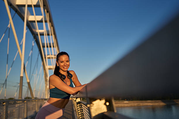 Young sporty woman with headphones around her neck is taking a break, leaning on a bridge railing and smiling at the camera during a beautiful sunset