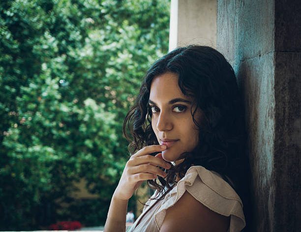 Very attractive young woman looking sideways a camara,lightly touching her lips dressed in a white suit, leaning against a marble wall and green background of a forest, copy space.