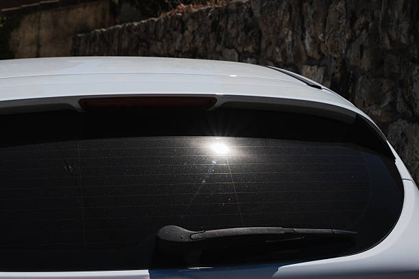 Sunlight reflects intensely off the rear window of a white car, highlighting the glass's heating lines and a rear wiper. The scene captures the interaction between solar energy and automotive technology in an urban environment. Ideal for topics related to renewable energy, vehicle efficiency, and modern automotive design.