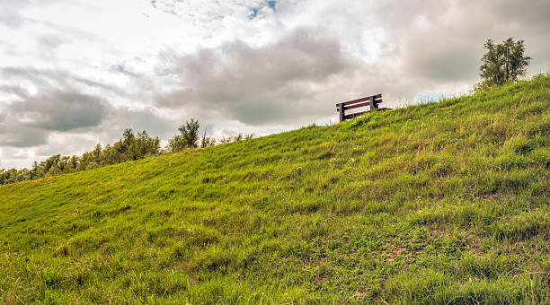 A simple wooden bench atop a Dutch dike. The bench is empty. The photo was taken on a cloudy day in summer.