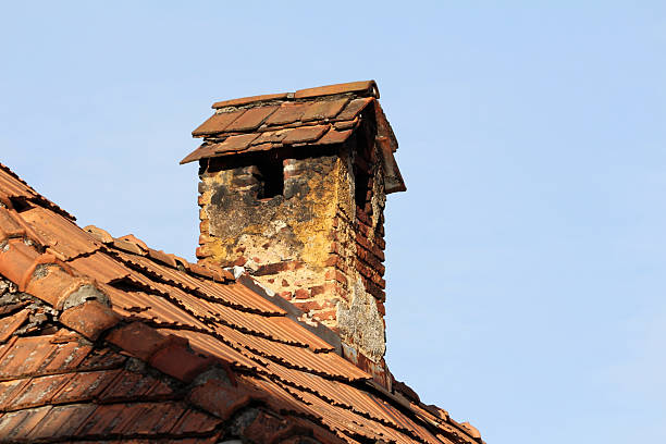 A brick chimney crowned with a tiled cap shows heavy soot stains and weathered mortar, rising from an old, crumbling terracotta roof against a clear blue sky