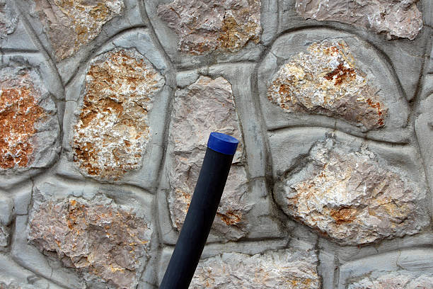 A black plastic water pipe with a blue cap protrudes diagonally from a decorative concrete wall made to mimic natural stone, used for utility access or irrigation systems