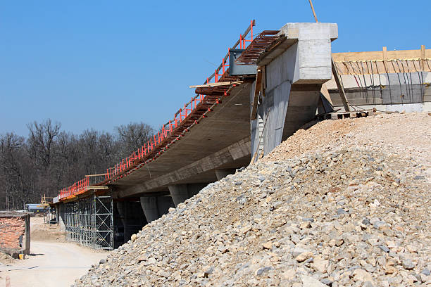 Partially built concrete bridge with visible rebar and scaffolding extends over a gravel mound, illustrating active civil engineering and large-scale infrastructure development