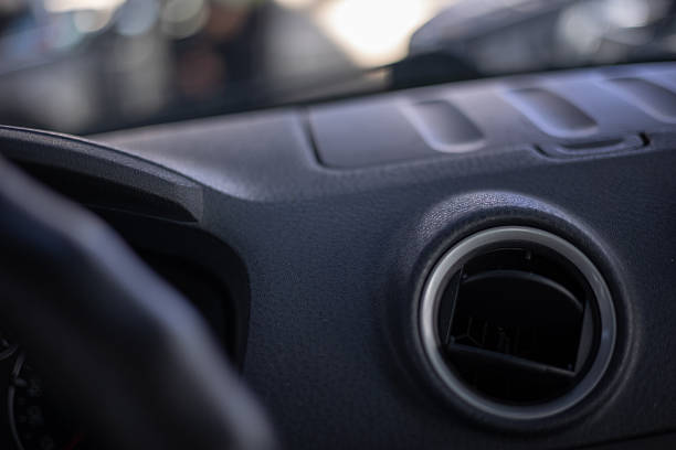istock A close-up shows a black plastic car dashboard with a round air vent featuring a chrome trim and horizontal louvers, with a blurred background and a partial view of the steering wheel on the left. 2227159127
