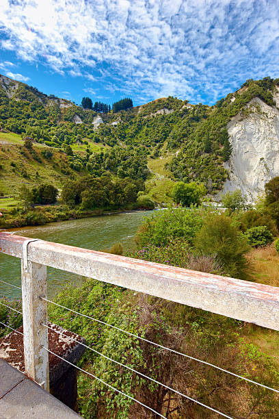 A picturesque landscape featuring a verdant river valley surrounded by forested cliffs and rugged terrain, accompanied by a rustic concrete railing in the foreground under a vibrant blue sky.