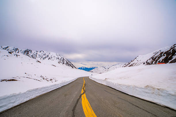 A paved road with a prominent double yellow line winds through a dramatic snow-covered mountain pass under a cloudy sky.