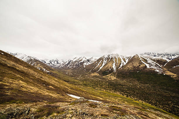 An expansive view showcases a vast mountain valley with rolling, rugged terrain leading up to snow-capped peaks under a cloudy sky.