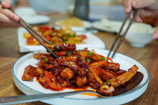 A close-up captures a vibrant dish of stir-fried eggplant with peppers and bean paste, served on a white plate. Hands holding chopsticks are reaching for portions of the food, conveying a shared Chinese dining experience in a restaurant setting.