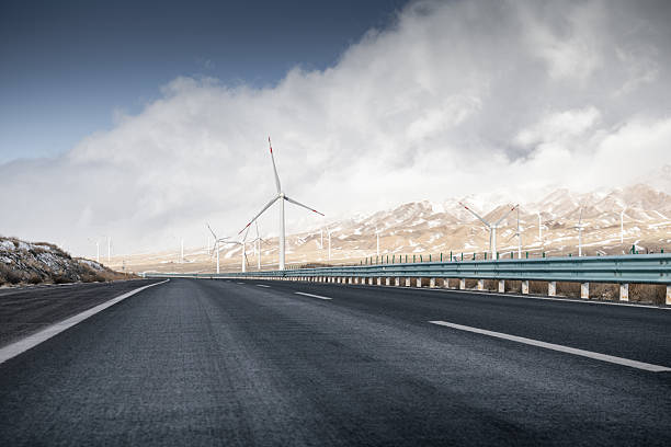 The image depicts a smooth asphalt road extending into the distance with wind turbines visible in the background. The scene includes a mountainous area with snow-capped peaks and a cloudy sky. The wind turbines are part of a renewable energy setup, contributing to sustainable power generation. The road is flanked by a guardrail, and the overall landscape showcases a blend of modern infrastructure and natural beauty.