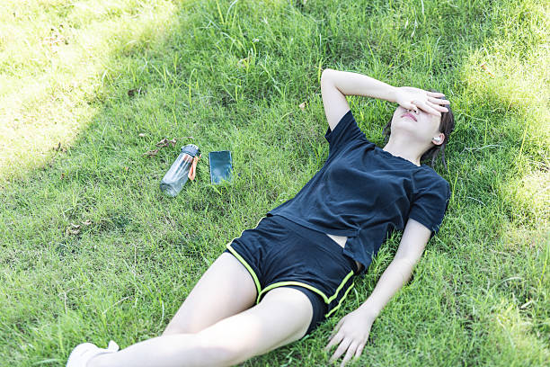 A young woman is lying on a green grassy area, resting with her hand covering her eyes. She is wearing a black t-shirt and black shorts with yellow trim, and white sneakers. There are a couple of plastic bottles nearby. She appears to be relaxing and taking a break in an outdoor, sunlit environment, enjoying a moment of leisure and stress relief.