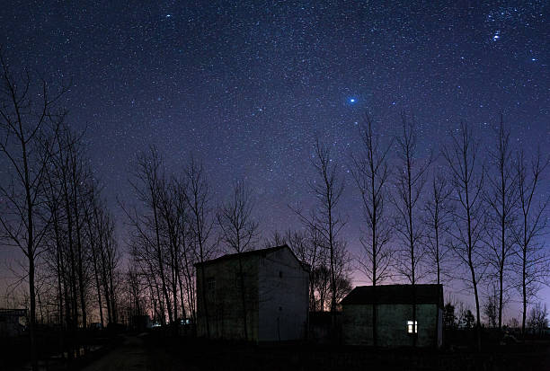 The image depicts a starry night sky above a quiet countryside area with bare trees and houses. The sky is filled with numerous stars, creating a beautiful and serene nighttime scene. The bare trees are silhouetted against the dark sky, and the houses are also in silhouette, adding to the peaceful and calm atmosphere of the rural landscape.