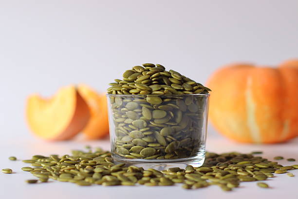 Glass jar filled with green pumpkin seeds or pepitas on a white surface, surrounded by scattered seeds