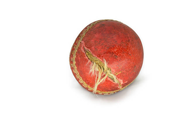Cut out of one old weathered run down ripped torn cut rough rustic cracked red maroon textured deformed frayed distorted leather cricket sports ball with seam and shadow isolated over white background.