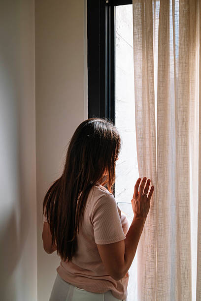istock A woman is looking out a window with a white curtain. 2227198000