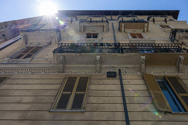 A striking upward view of a classic Italian apartment building basking in sunlight, featuring textured stucco walls, ornate iron balconies, and traditional wooden shutters on tall windows. Sun flares and a clear blue sky add warmth and vibrancy, evoking the charm of Italy’s architectural heritage and urban lifestyle. Perfect for illustrating southern European culture, Mediterranean city living, or travel concepts.