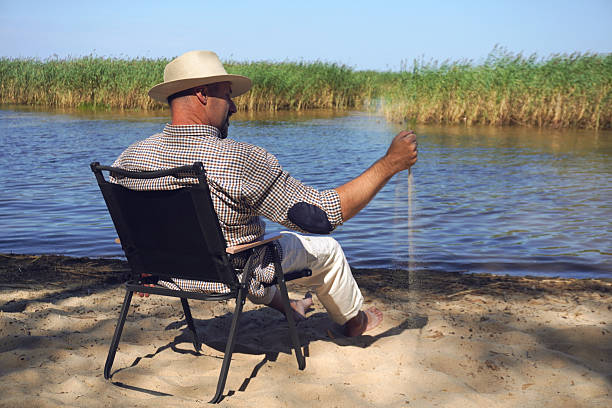 The man sits on a chair by the lake shore and fishes with a fishing rod. He is dressed in casual clothes. Outdoor recreation while fishing.