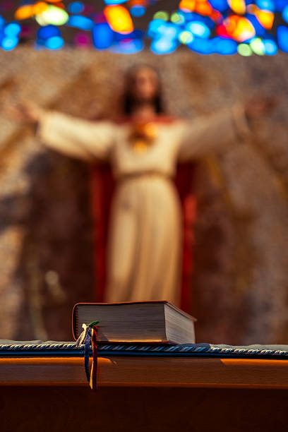 Close-up of a Bible on a church altar with a blurred statue of Jesus and colorful stained glass in the background.
