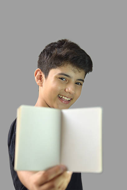 One cute smart happy ecstatic excited smiling laughing good looking boy holding showing a blank page or white paper label of open diary note pad notebook score card with copy space looking into the camera against gray background. Apt for advertisement, mock up posters banners.