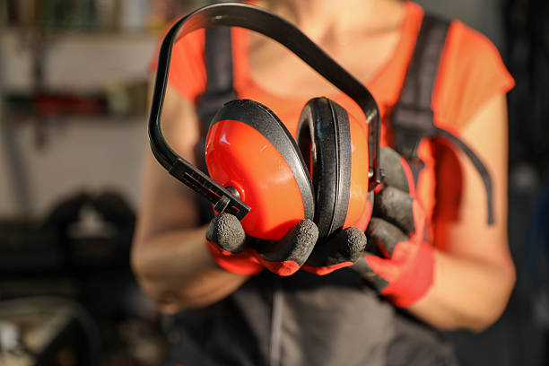 Close-Up Of A Female Worker In Protective Workwear Holding Red Noise-Canceling Headphones With Gloved Hands. The Image Emphasizes The Importance Of Safety Equipment In Industrial Settings, Showcasing Occupational Awareness, Precision, And Preparedness. Background Details Reinforce The Workshop Environment.