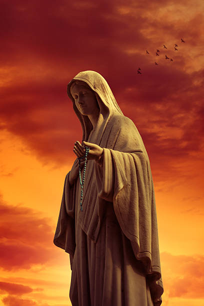 Statue of the Virgin Mary holding a rosary, silhouetted against a dramatic sunset sky with glowing cross in the background.