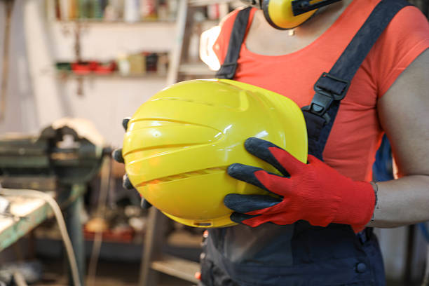 Close-Up Image Of A Female Worker Wearing Red Protective Gloves While Holding A Yellow Safety Helmet In A Workshop. The Focus On Hands And Gear Highlights Workplace Safety, Responsibility, And Professionalism In Industrial Environments. Tools And Equipment In The Background Add Authenticity To The Scene.