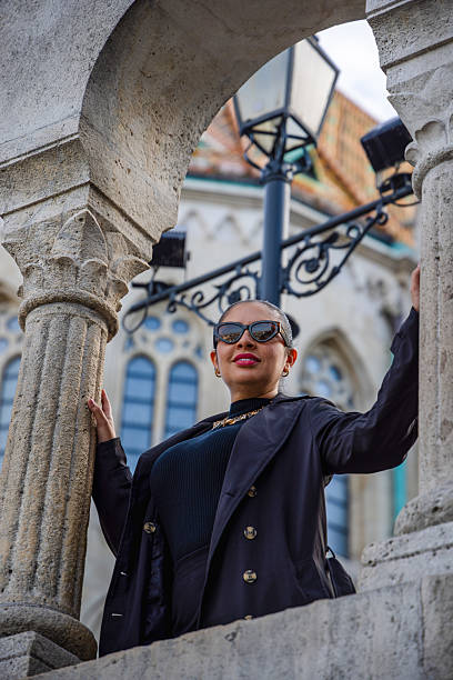 A stylish woman posing between ancient stone columns with a historical European building in the background. Captured from a low angle, this architectural portrait blends fashion and heritage.