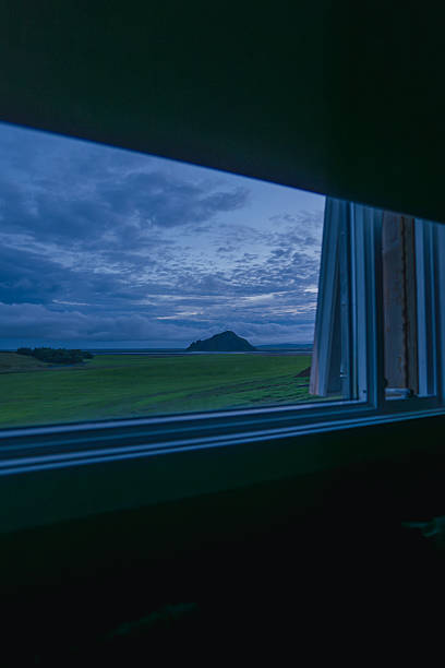 A serene grassy landscape at dusk is framed by an open window, with a distinct hill in the distance under a cloudy sky with soft blue tones.