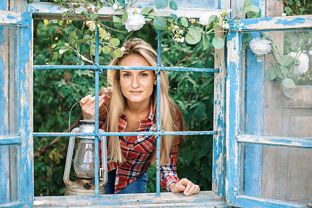 Young woman in plaid shirt looking through weathered blue window frame while holding an antique oil lamp in garden setting