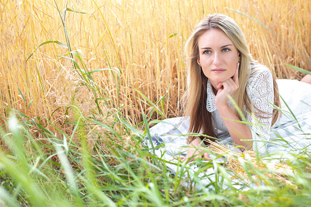 Beautiful blonde woman in white lace dress lying on picnic blanket surrounded by golden wheat stalks during sunset