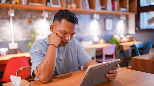 Young man is sitting at wooden table in cozy cafe, focused on tablet. He has thoughtful expression, resting his chin on his hand, surrounded by warm and inviting atmosphere