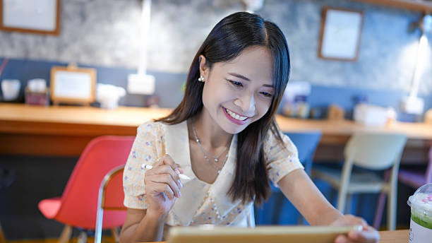 istock Young woman is sitting cafe, smiling while using tablet. She is dressed light colored blouse and appears engaged and happy. background features cozy atmosphere with wooden elements and soft 2227238493