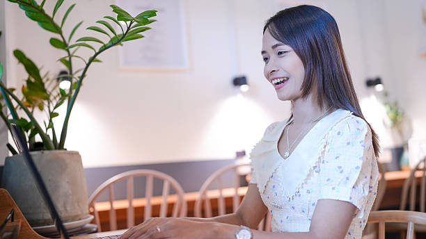 Young woman is smiling while working on laptop in cozy cafe setting. atmosphere is warm and inviting, with plants and soft lighting enhancing ambiance