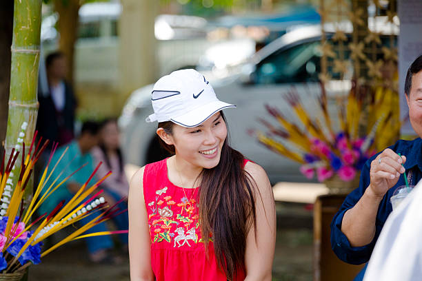Happy laughing young thai woman captured at public ceremony in park Kelang Nakhon Public Park in Lampang city in north Thailand. .Woman has long hair and is wearing a white cap and a red tank-top with flowers. She is standing near a cauldron with incense sticks. She is looking to her left side and is toothy laughing.  At right side 2 people are partly visible. People are attending public ceremony with monks and drums and drummers in park