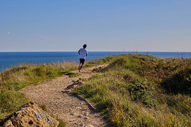 Man jogging on a coastal dirt trail with sea view under clear blue sky.