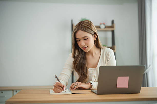 istock Young woman engaged in creative work at a modern desk, writing notes with a laptop in the bright and cozy workspace, focused on her tasks and ideas 2227247830