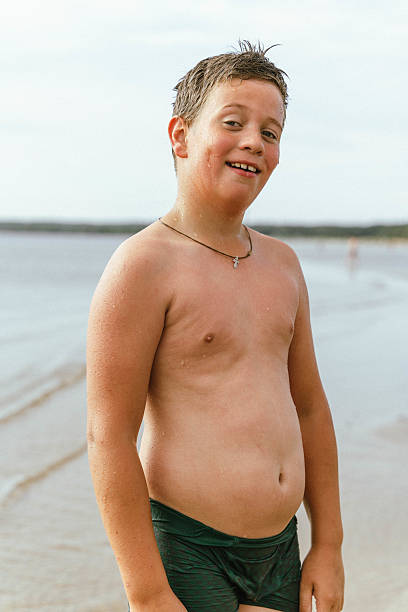 Smiling overweight boy wearing green swimming trunks and a cross necklace is enjoying summer vacation on a beach