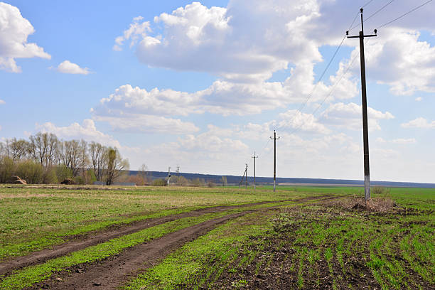 Dirt road through green field with power lines under cloudy sky