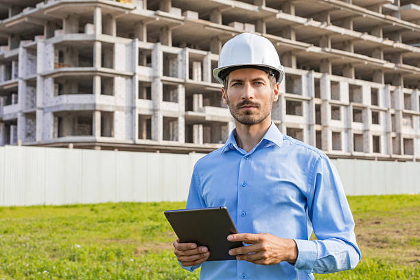 istock Handsome man with worker's helmet holding digital tablet while looking at camera, background buildings under construction. 2227272859