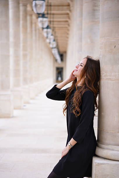 A fashionable young Middle Eastern woman with curly hair wearing sunglasses stands in front of the Eiffel Tower on a cloudy day.
