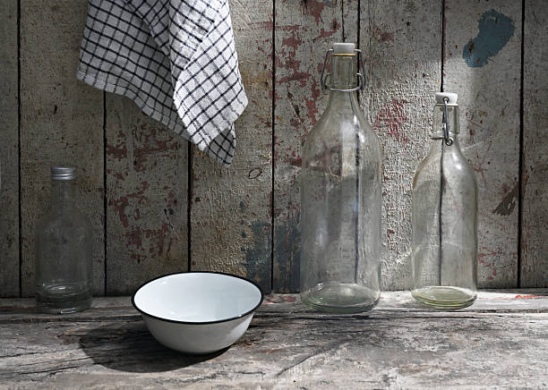 A rustic kitchen still life featuring an enamel bowl, three empty glass bottles, and a checkered dish towel hanging against a weathered wooden wall. The natural lighting and textured surfaces evoke a vintage farmhouse atmosphere, ideal for themes of sustainability, simplicity, and country living.