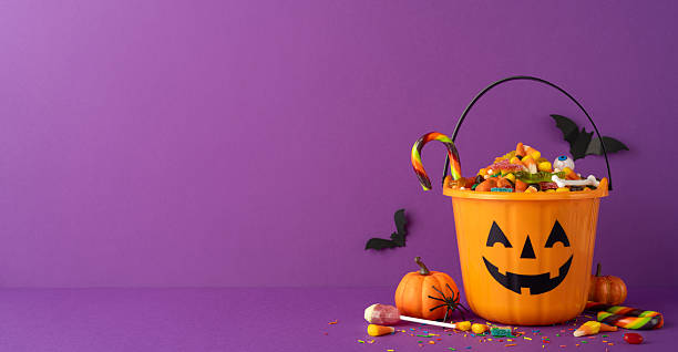 istock Orange jack-o-lantern bucket filled with assorted Halloween candy with a side view table purple backdrop 2227284188